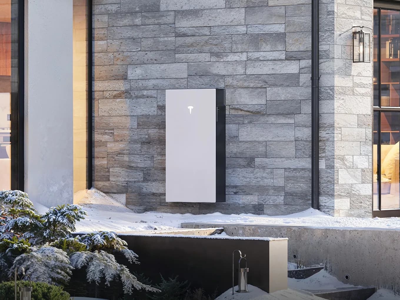 A white Tesla Powerwall 3 mounted on a stone wall outside a house in the snow, with warm lit windows either side